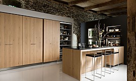 Optisch den Raum bestimmend ist das Eichenfurnier Sägerau, das durch seine Sägeschnittstruktur, die horizontal zu der vertikal ausgerichteten Maserung verläuft, der Oberfläche Robustheit verleiht und perfekt mit den kühlen Edelstahlgeräten harmoniert. Zuordnung: Stil Moderne Küchen, Planungsart Küchenzeile