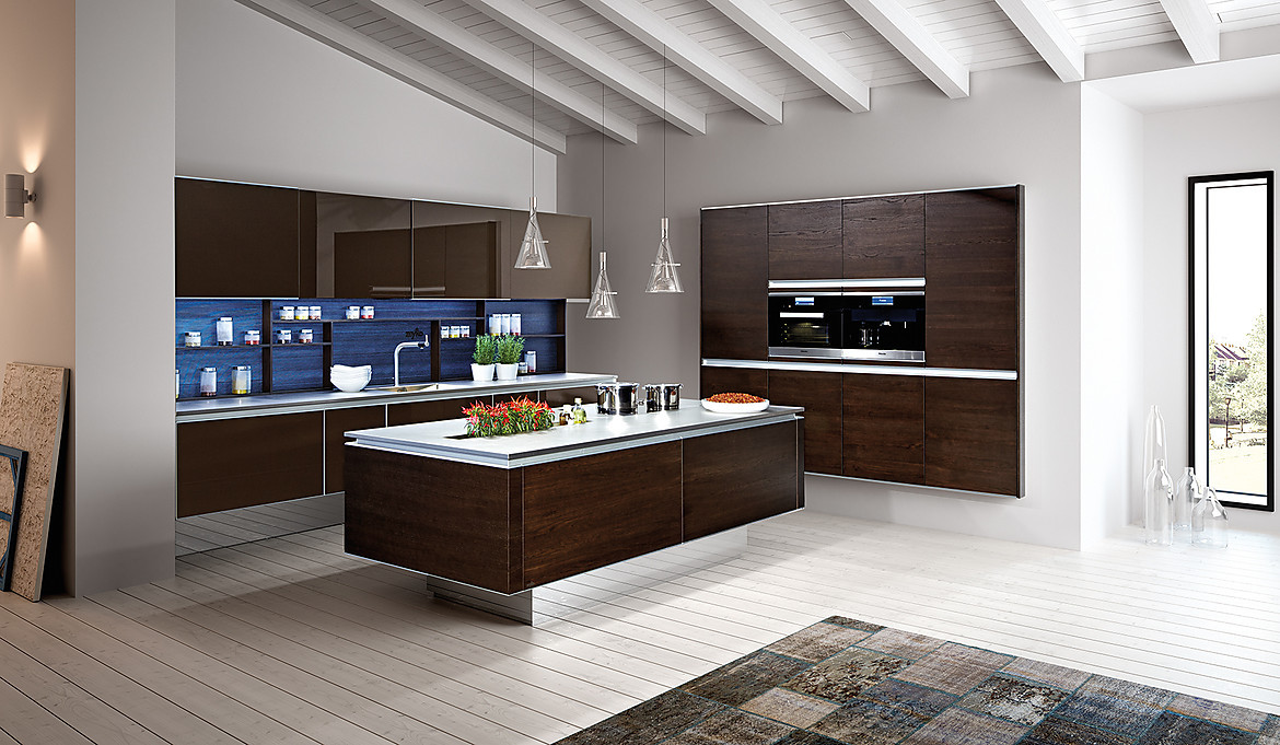 Elegante Inselküche mit dunklen grifflosen Fronten in massiver Eiche. Die Küchenrückwand erstrahlt in blauem Glas, was dem Gesamterscheinungsbild der Küche eine edle Note verleiht. Zuordnung: Stil Design-Küchen, Planungsart Küche mit Küchen-Insel