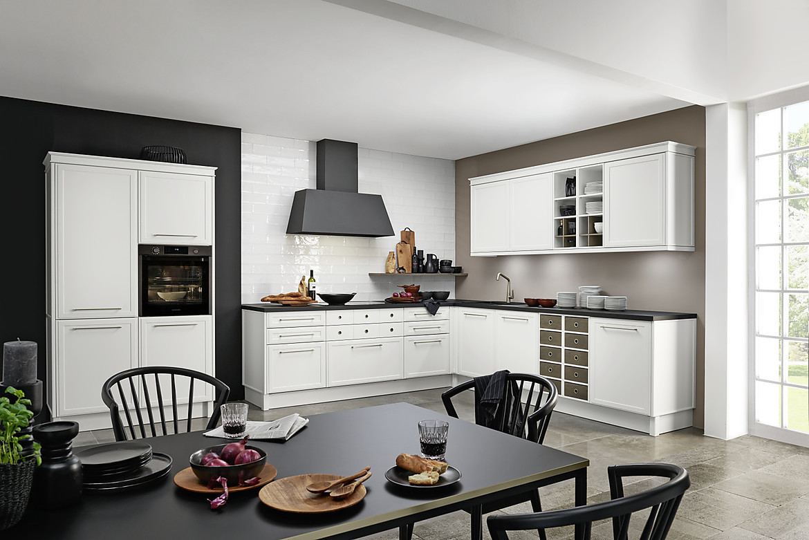 Beim Design der Küche handelt es sich um eine skandinavische Interpretation der Landhausküche. Die matten Fronten in Weiß wirken kühl und lassen zudem den Raum groß wirken. Die dunkle Arbeitsplatte und die Dunstabzugshaube setzen hier einen schönen Kontrast. Zuordnung: Stil Landhausküchen, Planungsart L-Form-Küche