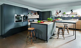 Beinahe modular wirkt dieses Beispiel einer urbanen Küchenlandschaft. Die grifflose Küche in Grau, Schwarz und mit Holzelementen besteht aus einer Kücheninsel mit Theke, Gerätehochschränken und einer Küchenzeile, in der die Spüle untergebracht ist. Holzregale und Tafellack unterstützen den Industrial Style. Zuordnung: Stil Design-Küchen, Planungsart Offene Küche (Wohnküche)